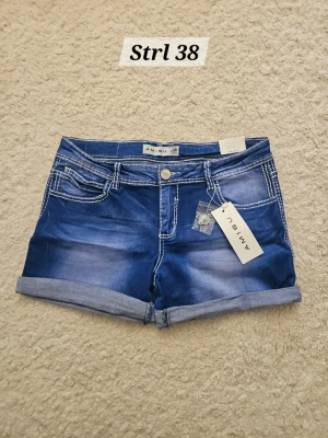 Blå denimshorts från Amisu - Nya! Snygga blå denimshorts från Amisu med uppvikta benslut och kontrastsömmar. Klassisk femficksmodell med knapp och dragkedja framtill. Perfekta för varma dagar och enkel att matcha med olika toppar.