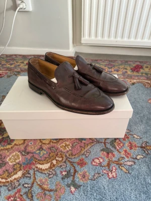 Brunello Cucinelli Tassel Loafers - Säljer ett par klassiska bruna loafers i läder med tofsar på ovansidan. Skorna har en stilren design med rundad tå och låg klack. Insidan är fodrad i ljust läder för extra komfort. Perfekta för dig som gillar tidlös och elegant stil.