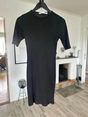 Svart ribbad klänning 157 M - En svart ribbad klänning från 157 med kort ärm och rund halsringning. Modellen är kroppsnära och har en enkel, stilren design som passar perfekt till sneakers eller boots. Materialet är stretchigt och bekvämt.