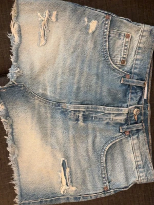 Mini kjol  - Säljer en ljusblå jeans kjol med råa kanter och slitna detaljer både fram och bak. Kjolen har klassisk femficksdesign, bälteshällor och knappgylf. Perfekta för en avslappnad och trendig sommarstil.
