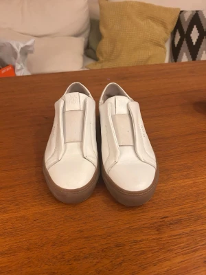 Vita sneakers från Axel Arigato - Stilrena vita slip-on sneakers från Axel Arigato med minimalistisk design. Skorna har ovandel i slätt läder och en tjock, ljusbrun gummisula. Diskret logga på sidan och plös. Perfekta för dig som gillar enkel och clean stil. Väldigt sparsamt använda då de är för små. Är öppen för att diskutera pris💕