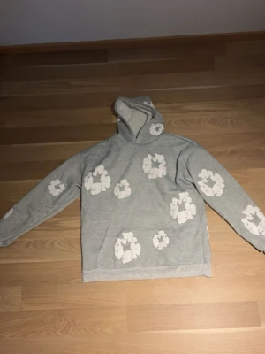 Grå hoodie med vita blommor - Säljer en grå hoodie med stora vita blommönster över hela tröjan. Hoodien har huva och en klassisk känguruficka framtill. Mjuk och skön bomullsmix som passar perfekt till chill dagar. Oversized fit för en avslappnad stil.
