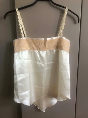 Beige och gräddvit linnetopp med spets - Söt linnetopp i beige och gräddvitt med spetsdetaljer upptill och broderade blommor på axelbanden. Toppen har ett glansigt material och tunna axelband i virkad stil. Perfekt för en somrig och romantisk look. Armhåla till armhåla 40 cm