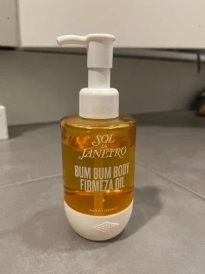 Sol de Janeiro Bum Bum Body Oil - Sol de Janeiro Bum Bum Body Firmeza Oil i en genomskinlig pumpflaska med vit topp och botten. Oljan är gyllengul och flaskan rymmer 100 ml. Perfekt för dig som vill ha en lyxig kroppsolja med brasiliansk inspiration.