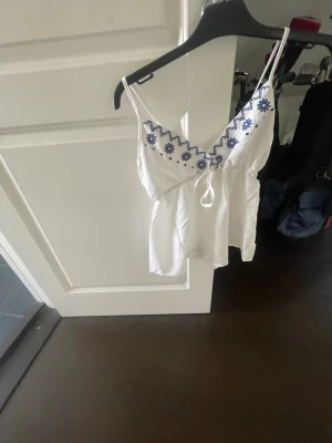 Vit broderad linnetopp med knyt - Supersöt vit linnetopp med tunna axelband och blå broderade blommor längs v-ringningen. Toppen har ett litet knyt framtill och luftig passform, perfekt för varma dagar❤️