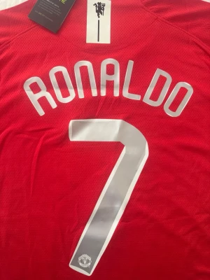 Manchester United Ronaldo tröja - Röd fotbollströja från Manchester United med Ronaldo och nummer 7 tryckt i vitt på ryggen. Tröjan har ventilerande meshmaterial och MUFC-detalj i nacken. Perfekt för dig som älskar fotboll och vill bära din idol på ryggen.