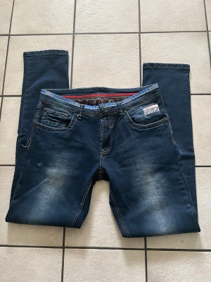Mörkblå slim fit jeans med slitningar - Snygga mörkblå jeans i slim fit-modell med diskreta slitningar på låren och knäna. Klassisk femficksdesign, kontrastsömmar och detaljerad insida i midjan med röd rand. Märke: Fashion. Perfekta för en trendig och avslappnad look.