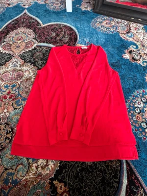 Röd blus med spetsdetalj - Snygg röd blus med långärmad design och dekorativ spets vid halsringningen. Blusen har knappar vid ärmsluten och en lätt v-ringad hals. Perfekt för dig som vill ha en färgstark och trendig look.