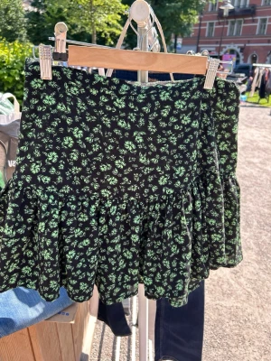 Svart blommig volangkjol Gina Tricot - Kort svart kjol från Gina Tricot med grönt blommönster och volang nedtill. Kjolen har en skön passform och elastisk midja, perfekt för dig som gillar en söt och trendig stil. Lätt material som passar bra till varma dagar.