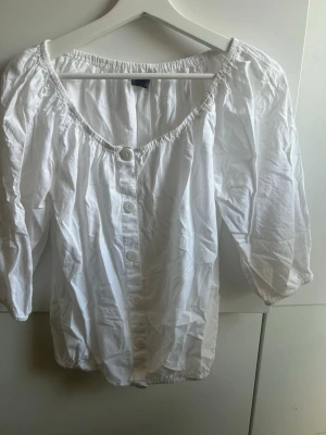 Vit offshoulder blus med knappar - Snygg vit offshoulder blus med knäppning framtill och trekvartsärmar. Blusen har resår vid halsringning, ärmslut och nederkant för en avslappnad passform. Perfekt till jeans eller kjol för en trendig look.