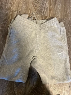 Grå sweatshorts från Nike - Mysiga grå sweatshorts från Nike med snörning i midjan och två sidofickor. Klassisk Nike-logga broderad på vänster ben. Perfekta för chill dagar eller träning. Tillverkade i mjuk bomullsblandning för maximal komfort.