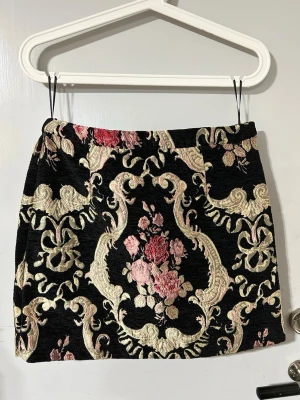 Broderad kortkjol från Bik Bok - Säljer en svart kortkjol från Bik Bok med broderade blommor i rosa och beige toner. Kjolen har ett elegant, barockinspirerat mönster och är tillverkad i ett tjockare tyg som ger en lyxig känsla. Perfekt för dig som vill sticka ut med en unik look.