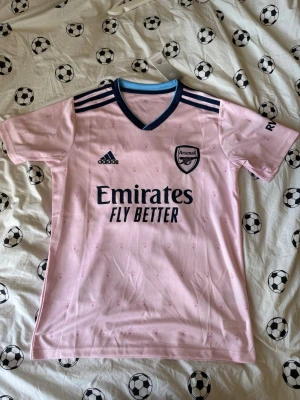 Arsenal rosa matchtröja Adidas - Snygg rosa Arsenal matchtröja från Adidas med mörkblå detaljer och klubbmärke på bröstet. Tröjan har korta ärmar, V-ringning och tre klassiska Adidas-ränder på axlarna. Tillverkad i lätt och ventilerande funktionsmaterial, perfekt för fotbollsträning.