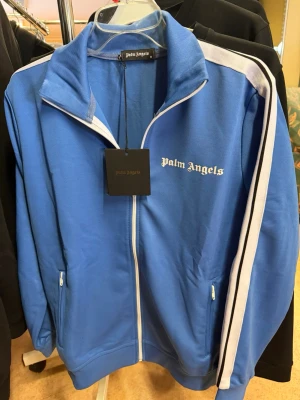 Blå track jacket från Palm Angels - Snygg blå track jacket från Palm Angels med vita ränder längs ärmarna och vit dragkedja. Jackan har ståkrage, två fickor med dragkedja och logga på bröstet. Perfekt för en sportig och trendig look. Materialet är mjukt och bekvämt.