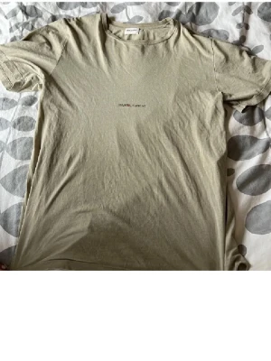 Beige Saint Laurent t-shirt - Snygg beige t-shirt från Saint Laurent med diskret logga broderad på bröstet. Klassisk passform med rund hals och korta ärmar. Perfekt för en stilren och avslappnad look.
