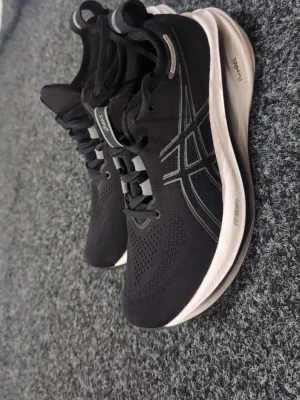 Svarta Asics sneakers med vit sula - Snygga svarta sneakers från Asics med klassisk logga på sidan och vit, tjock sula. Ovandelen är i mesh för bra andningsförmåga och skorna har svarta snören. Perfekta för dig som gillar sportig stil och komfort.