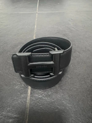 Svart bälte med H-spänne - Snyggt svart bälte från med ikoniskt H-format spänne i matt finish. Bältet är tillverkat i skinn och har en stilren, minimalistisk design som passar till många olika outfits. Perfekt accessoar för dig som gillar exklusiva detaljer.