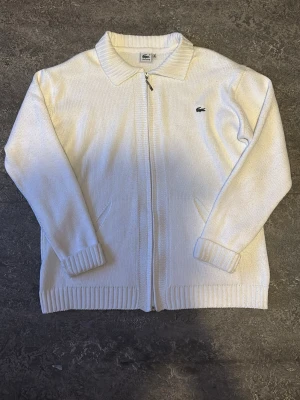 Vit ziptröja från Lacoste - Klassisk vit stickad ziptröja från Lacoste med krage och dragkedja framtill. Tröjan har ribbade muddar, två fickor och den ikoniska gröna krokodilloggan broderad på bröstet. Perfekt för en clean och stilren look.