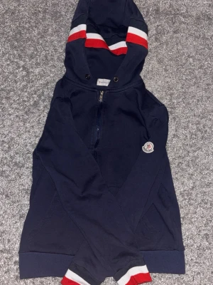 Mörkblå Moncler hoodie med dragkedja - Snygg mörkblå hoodie från Moncler med dragkedja framtill och klassisk Moncler-logga på ärmen. Huva med snörning och ribbade muddar i rött, vitt och svart. Perfekt för en avslappnad och trendig look. Tillverkad i mjukt bomullsmaterial.