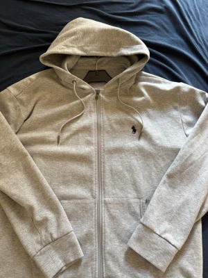Grå zip hoodie från Polo Ralph Lauren - Snygg grå hoodie med dragkedja från Polo Ralph Lauren. Klassisk huva med dragsko, två fickor framtill och ikoniska broderade loggan på bröstet. Tillverkad i mjuk bomullsblandning som känns skön mot huden. Perfekt för en avslappnad och stilren look.