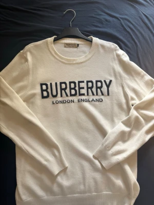 Vit stickad tröja från Burberry - Snygg vit stickad tröja från Burberry med svart text 'BURBERRY LONDON ENGLAND' på bröstet. Tröjan har rund halsringning och långa ärmar. Perfekt för dig som gillar klassisk och stilren design. (Står XL men sitter som en M)