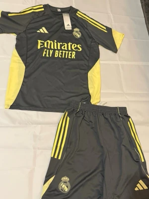Real Madrid Barcelona matchställ Adidas - Två Real Madrid matchställ från Adidas. Ett set är mörkgrått med gula detaljer och ett är vitt med svarta och lila inslag. Båda seten har klubbmärke, Adidas-logga och 'Emirates Fly Better' tryck på tröjan. Tillverkade i lätt och ventilerande material, perfekt för fotbollsträning. Och en Barcelona 