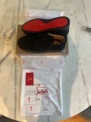 Svarta sneakers från Christian Louboutin - Snygga svarta sneakers från Christian Louboutin med den ikoniska röda sulan. Skorna har en klassisk låg profil, svarta skosnören och ovandel i textil. Perfekta för dig som vill ha en stilren och exklusiv look med en touch av lyx.