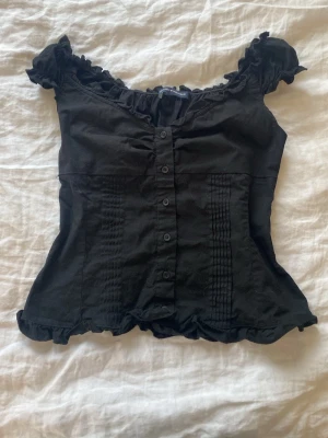 Svart blus med volang från Brandy Melville - Supersöt svart blus från Brandy Melville med knappar framtill, volangkant vid ärmar och nederkant samt lätt rynk vid bysten. Blusen har korta puffärmar och en figurnära passform💕