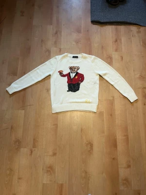 Vit stickad tröja Polo Ralph Lauren - Vit stickad tröja från Polo Ralph Lauren med ikonisk björn i smoking och röd kavaj på bröstet. Rund halsringning och ribbstickade muddar. Perfekt för dig som vill ha en unik och klassisk look med en twist.