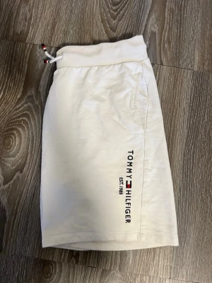 Vita shorts från Tommy Hilfiger - Snygga vita shorts från Tommy Hilfiger med broderad logga och text på ena benet. Shortsen har snörning i midjan och en bakficka med liten logga. Perfekta för sommardagar och har en clean, stilren look.