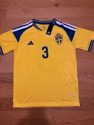 Sverige matchtröja Lindelöf #3 Adidas - Gul och blå officiell Sverige matchtröja från Adidas med Lindelöf och nummer 3 på ryggen. Tröjan har korta ärmar, broderat landslagsemblem och Climalite-material som andas. Perfekt för fotbollsträning eller att bära på läktaren.