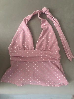Rosa prickig halterneck topp - Söt rosa halterneck topp med vita prickar och knyt i nacken. Toppen har djup v-ringning och är tillverkad i ett mjukt, stretchigt material som känns bekvämt mot huden. Perfekt för dig som vill ha en feminin och trendig look.