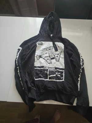 Svart Star Wars hoodie med tryck - Svart hoodie med  Star Wars-tryck på framsidan som visar Millennium Falcon och andra rymdskepp. Huva och tryck på ärmarna. 