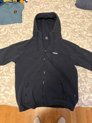 Mörkblå hoodie med dragkedja Djurgårn - Mörkblå hoodie från Djurgårn med dragkedja framtill, två fickor med dragkedja och justerbar huva. Märket sitter broderat på bröstet. Hoodien har en avslappnad passform och är tillverkad i mjukt material, perfekt för sena sommarkvällar 