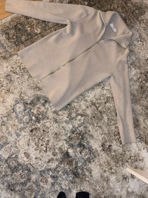 Beige ribbad ziptröja från LUNG - Säljer en beige ribbad ziptröja från LUNG med hög krage och hel dragkedja framtill. Tröjan har lång ärm och är tillverkad i ett mjukt, stretchigt material som ger en clean och stilren look.