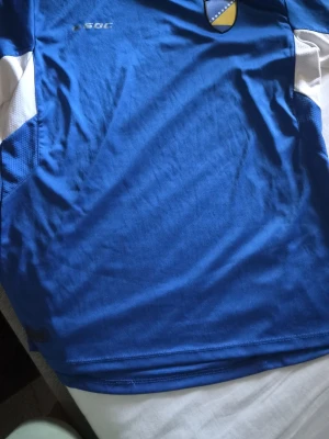 Blå fotbollströja från SOC - Snygg blå fotbollströja från SOC med vita detaljer på sidorna och ett tryckt emblem på bröstet. BOSNIEN.Tillverkad i ett lätt och snabbtorkande material som passar perfekt till träning eller match. Klassisk sportig design med rund hals.