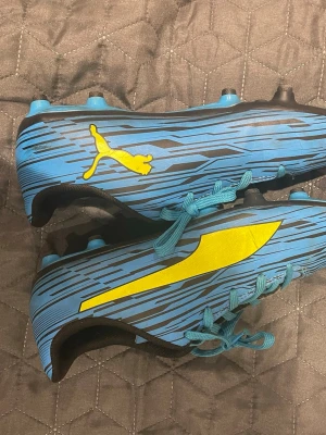 Blåa Puma fotbollsskor med gult tryck och vit puma med orange tryck - Säljer ett par snygga blåa fotbollsskor från Puma med coolt mönster och gula detaljer. Skorna har snörning, låg profil och dobbar i plast för bra grepp på planen. Perfekta för dig som vill sticka ut på matchen!