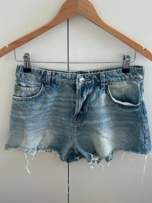 Blå jeansshorts med fransig kant - Snygga blå jeansshorts med rå, fransig kant och klassisk femficksdesign. Shortsen har en avslappnad passform och är perfekta för varma dagar. Tillverkade i denim med slitna detaljer för en cool look.