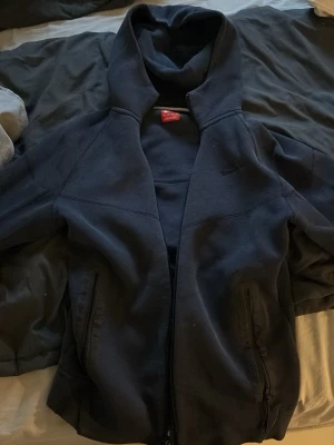 Svart Nike tech hoodie med dragkedja - Säljer en mörkblå Nike tech hoodie med dragkedja framtill och två fickor med dragkedja. Hoodien har en stor huva och är tillverkad i mjukt sweatshirtmaterial. Diskret Nike-logga på bröstet. Perfekt för en avslappnad och sportig stil.