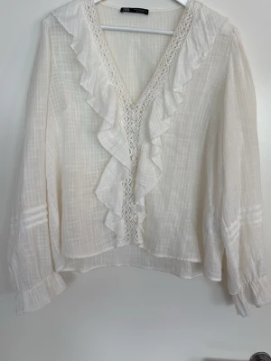 Vit blus med volanger från Zara - Romantisk vit blus från Zara med djup V-ringning, volangdetaljer och spets längs halsen. Tillverkad i ett tunt, lätt och något transparent tyg med långärmade ärmar och diskreta rynkade muddar. Perfekt för en chic och avslappnad look.