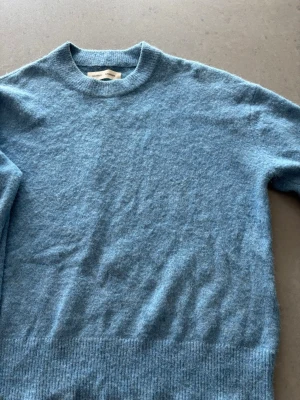 Blå stickad tröja från Samsøe Samsøe - Mysig blå stickad tröja från Samsøe Samsøe med rund halsringning och ribbade muddar. Tröjan har en enkel och stilren design, perfekt för lager på lager. Materialet ser ut att vara mjukt och fluffigt, troligen ull eller en ullmix. Något lite ljusare/ skavt på baksida ärm vid armbågen.  Annars jättefin!