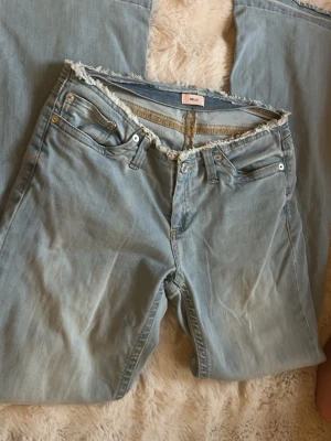 Ljusblå jeans med rå kant från Nelly - Säljer ett par ljusblå jeans från Nelly med snygg rå kant upptill. Klassisk femficksmodell med dragkedja och knapp. Jeansen har en avslappnad passform och är tillverkade i denim med slitna detaljer för en cool look.