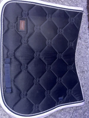 Marinblått schabrak Equestrian Stockholm - marinblått schabrak från Equestrian Stockholm med quiltat mönster och vit kant. Schabraket har en glansig yta och broderad logga i tyget. Använt fåtal gånger finns inga skador på de lite dammigt på bilden då de legat nedpackat men tvättas självklart innan jag postar.