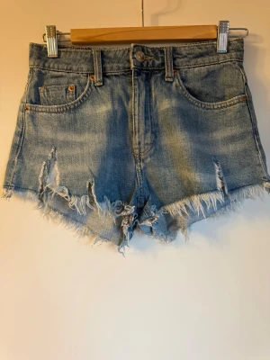 Blå jeansshorts med fransig kant - Snygga blå jeansshorts med rå, fransig kant och slitna detaljer framtill. Klassisk femficksmodell med knapp och dragkedja. Perfekta för varma dagar och ger en avslappnad vibe.