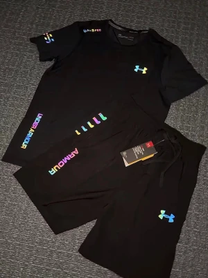 Svart träningsset från Under Armour - Snyggt svart träningsset från Under Armour med t-shirt och shorts. Setet har färgglada loggor och detaljer i regnbågsfärger. Perfekt för gymmet eller löprundan. Materialet är lätt och andas, vilket gör det bekvämt under träning. Storlek M helt ny 