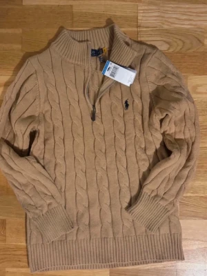 Beige kabelstickad tröja Polo Ralph Lauren - Kabelstickad beige tröja från Polo Ralph Lauren med halv dragkedja och ribbade muddar. Tröjan har en hög krage och den klassiska mörkblå logotypen broderad på bröstet. Perfekt för dig som gillar stilren och tidlös design.