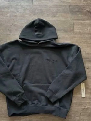 Svart Essentials hoodie - Svart hoodie från Essentials med diskret logga på bröstet. Klassisk modell med huva och känguruficka framtill. Tillverkad i mjuk bomullsblandning som är skön att bära. Perfekt för en avslappnad och stilren look.