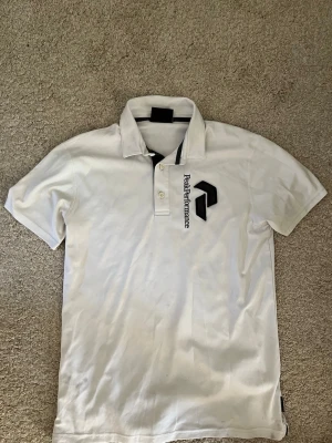 Vit Peak Performance pikétröja - Klassisk vit pikétröja från Peak Performance med svart logga och text på bröstet. Tröjan har korta ärmar, krage och knappar framtill. Perfekt till en avslappnad men ändå stilren look. Skönt sport aktigt material! Storlek L på tröjan men passar perfekt på M!
