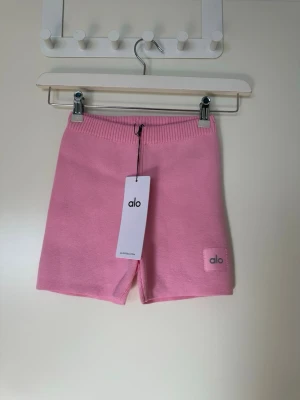 Rosa stickade shorts från Alo - Säljer ett par ljusrosa stickade shorts från Alo. De har en ribbad midja och en liten Alo-logga på benet. Perfekta för en avslappnad och trendig look. Supersköna och mjuka i materialet.