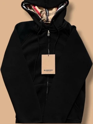 Burberry zip - Snygg svart hoodie från Burberry med dragkedja och klassisk huva. Insidan av huvan har Burberrys ikoniska rutmönster och detaljer i beige. Hoodien har fickor på sidorna och guldfärgade metalldetaljer på snörena. Perfekt för en stilren och avslappnad look. Storlek S